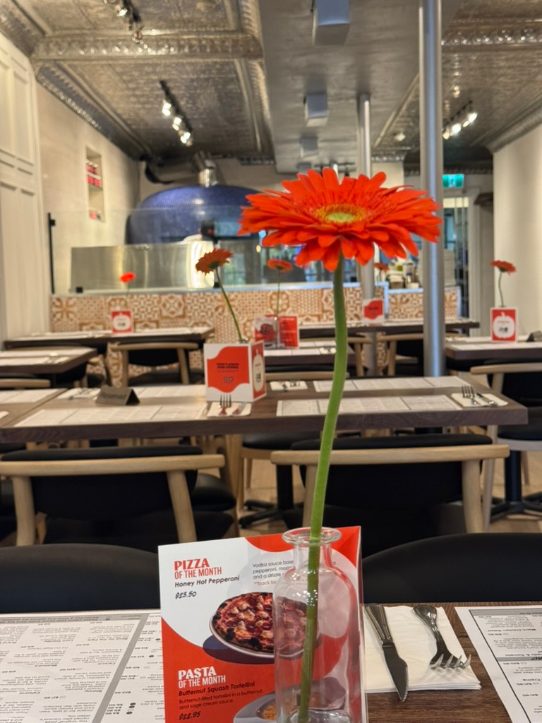 A flower on a set table inside the Bloor West Village dining room, with the blue pizza oven beyond